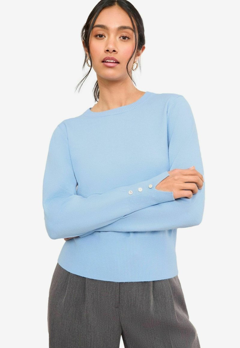 Ljusblå stickad tröja med rund halsringning, ribbat avslut och tre knappdekorationer på mudden. Slät textur, åtsittande design.