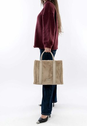 Vrouw draagt een bordeauxrode trui en jeans, houdt een beige nepbont totebag met leren handvatten vast, staand tegen een witte achtergrond.