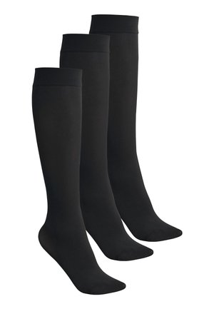 Jambes portant des chaussettes de compression noires montantes jusqu'aux genoux, debout sur la pointe des pieds sur un fond uni.