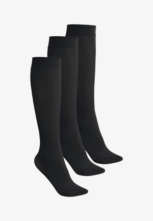 Jambes portant des chaussettes de compression noires montantes jusqu'aux genoux, debout sur la pointe des pieds sur un fond uni.