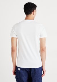 T-shirt en coton blanc à manches courtes, col rond, texture lisse, style ajusté, vu de l'arrière, porté avec un pantalon bleu.