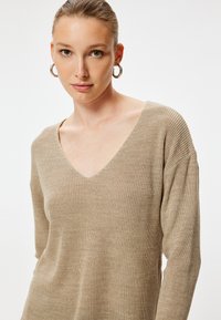 Beige gestrickter Pullover mit tiefem V-Ausschnitt, langen Ärmeln und strukturiertem Stoff, der locker sitzt und subtile Rippdetails aufweist.