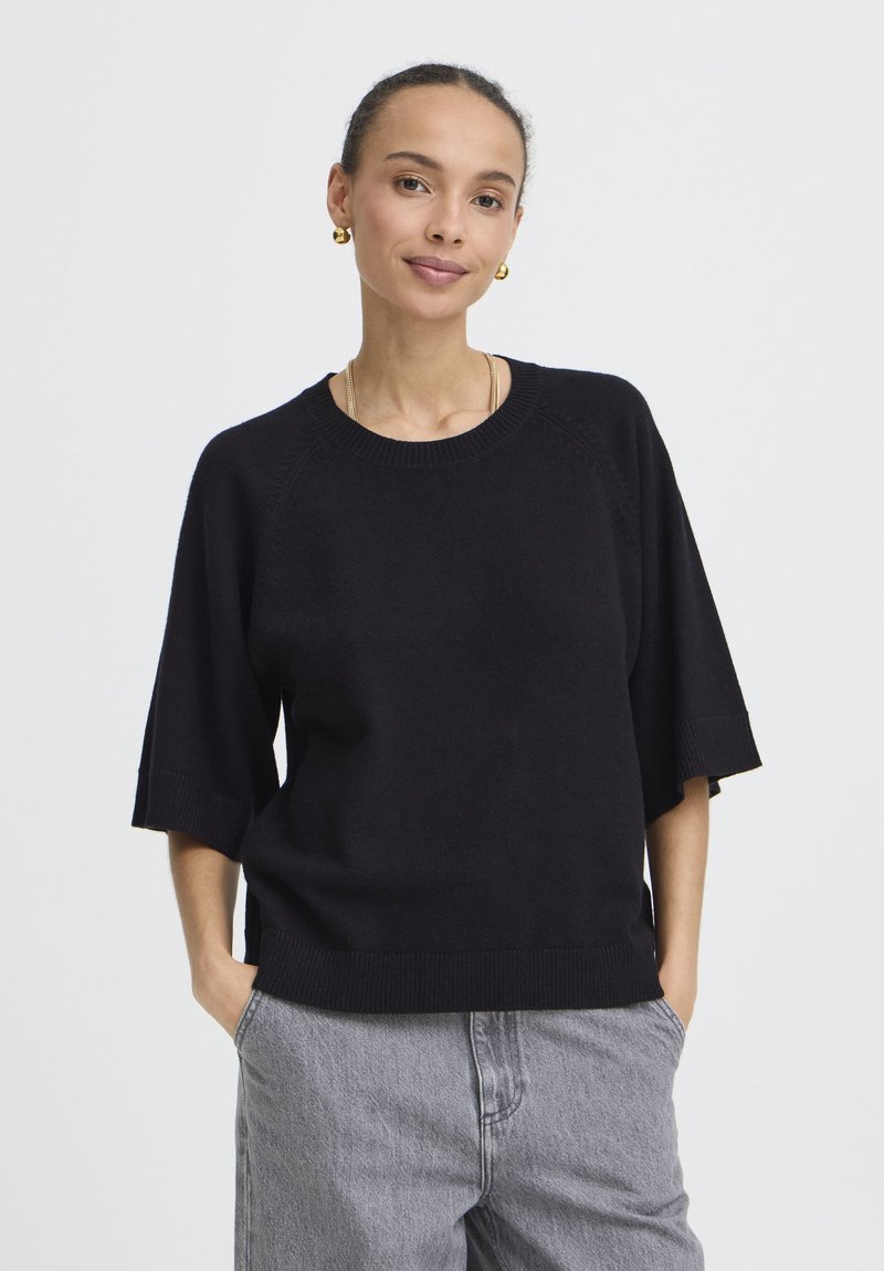 Femme debout les mains dans les poches, portant un pull noir à manches courtes, un pantalon gris, des boucles d'oreilles en or et un collier discret sur fond blanc.