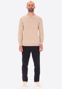 Uomo in piedi di fronte, indossa un maglione beige, pantaloni neri e sneakers bianche contro uno sfondo chiaro e semplice.