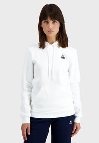 Hoodie blanc avec une poche frontale, capuche à cordon, et logo brodé sur la poitrine. Tissu doux et manches ajustées.