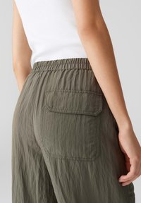 Lichte, olijfgroene broek met een ontspannen pasvorm, elastische tailleband en een enkele achterzak met een klep. Getextureerde stof zichtbaar.