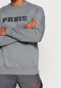 Felpa in pile grigia con il testo "PARIS" ricamato in nero. Presenta un collo rotondo e il logo rosso Nike sulla manica sinistra.