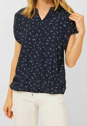 Vrouw draagt een donkerblauwe korte mouwen blouse met kleine witte bladvormen en witte broek, houdt met één hand een kraag vast.