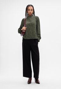 Πράσινη μπλούζα με ανθισμένο σχέδιο και ψηλό γιακά, σε συνδυασμό με μαύρο παντελόνι με φαρδιές γραμμές. Το μοντέλο κρατά μια μπορντό τσάντα χιαστί.