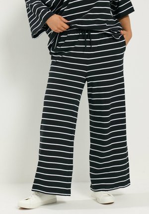 Personne portant un pantalon noir à jambes larges avec des rayures horizontales blanches et des baskets blanches, mains dans les poches et tenant l'ourlet de la chemise.
