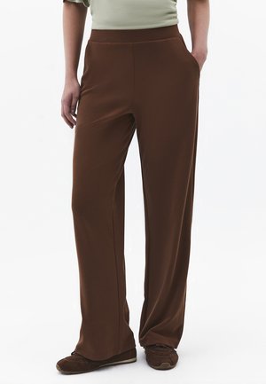 Person iført løse brune bukser med sidelommer og brune sneakers, mod en ensfarvet hvid baggrund.