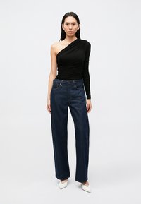 Top preto de um ombro só feito de tecido franzido, combinado com jeans escuros de cintura alta e sapatos brancos de salto fino e pontiagudo. Design simples e limpo.