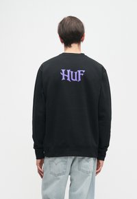 Pessoa vestindo um moletom preto com texto roxo "HUF" na parte superior das costas e jeans azul claro, de pé contra um fundo branco simples.