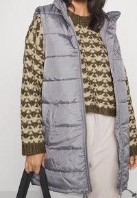 Žena v šedé prošívané bezrukávové bundě přes hnědou a béžovou vzorovanou pletenou svetr, držící černou tašku za rukojeť.