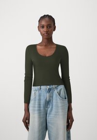 Mørkegrønn langermet topp med dyp V-hals. Det teksturerte stoffet sitter tettsittende på kroppen, sammen med lyseblå, løse jeans.