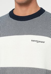 Pulover tricotat cu dungi albastre și albe, cu guler ribat. Prezintă logo-ul brodat "tommyjeans" pe piept. Material texturat.