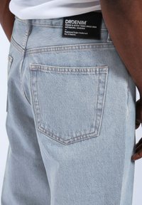 Jasnoniebieskie dżinsy o wyblakłej fakturze, z naszywką na pasku i pojedynczą tylną kieszenią z kontrastowym przeszyciem.