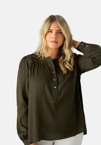Chemisier vert foncé à boutons avec un design à épaules plissées, manches longues et un tissu doux et texturé. Présente de petits boutons le long de la patte de boutonnage.