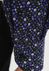 Koszula z guzikami na ciemnym tle, ozdobiona małymi fioletowymi i białymi wzorami kwiatowymi. Miękka tkanina z kontrastującymi czarnymi detalami guzików.