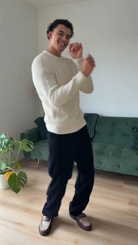 Una persona con un maglione lavorato a maglia color crema e pantaloni scuri danza vicino a un divano verde, con una pianta in vaso e pavimento in legno chiaro visibili.