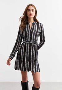 Zwarte button-up jurk met verticale witte strepen, lange mouwen, een ceintuur rond de taille en een relaxte pasvorm. Gladde stof met een gepolijste glans.