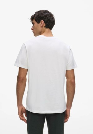 Homme aux cheveux courts et bouclés de dos, portant un t-shirt blanc uni à manches courtes et un pantalon foncé sur fond blanc.