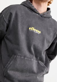Sudadera gris con capucha, con bolsillo tipo canguro y logo amarillo "ellesse Resort" en el frente. Textura suave y ligeramente desgastada.