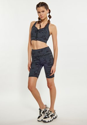 Tânără stând în bustieră sport neagră camuflaj și pantaloni scurți de ciclism cu talie înaltă asortați, cu adidași groși albi și negri.