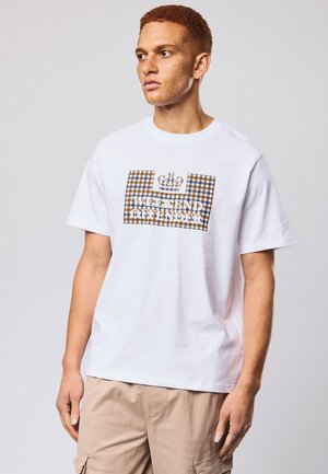 Ung mann med rødt hår iført hvit T-skjorte med rutemønster og teksten "Weekend Offender", samt beige cargobukser, som ser mot høyre.