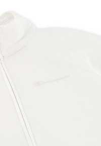 Giacca bianca con zip realizzata in tessuto morbido, dotata di colletto alto e logo goffrato discreto sul lato sinistro. Sono visibili i dettagli delle cuciture.