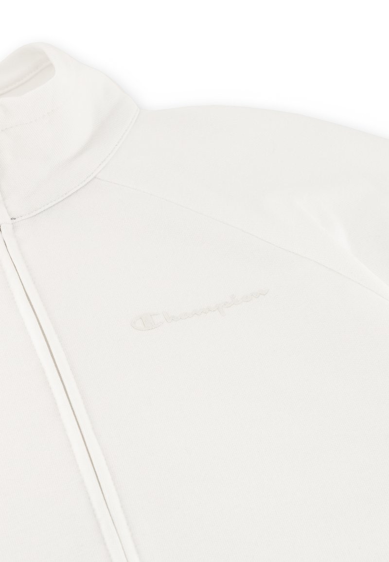 Weiße Zip-Jacke aus weichem Material, mit einem hohen Kragen und dezentem eingeprägtem Logo auf der linken Seite. Nahtdetails sind sichtbar.