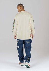 Beige Langarmshirt mit Druck auf den Ärmeln, kombiniert mit blauen Jeans mit gestickten Details und weißen Sneakers mit grünen Sohlen.