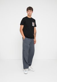 Black cotton t-shirt with a front graphic pocket design, paired with loose-fitting grey trousers and white sneakers.