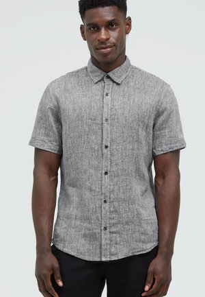 Mann trägt ein graues kurzärmeliges Hemd mit Knöpfen, steht vor einem einfarbigen hellen Hintergrund und blickt neutral nach vorne.