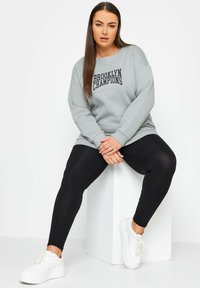 Grå sweatshirt med tryck av "BROOKLYN CHAMPIONS", kombinerad med svarta leggings och vita sneakers. Mjuk tyg, avslappnad passform och rundad halsringning.