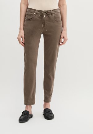 Vrouw draagt bruine slim-fit broek met trekkoord in de taille en zwarte instap loafers, staande tegen een effen lichte achtergrond.