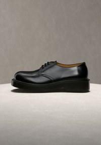 Chaussure habillée en cuir noir à lacets avec une semelle épaisse, présentée sur un fond beige neutre sur une surface claire.