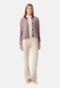 Court cardigan bordeaux avec un motif en losanges, boutons noirs, porté sur un pull beige et un pantalon beige ajusté, associé à des baskets claires.