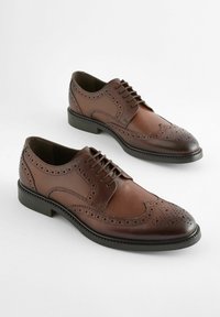 Chaussures derby en cuir marron avec un design brogue, bout pointu et semelle en caoutchouc plate. Perforations intriquées le long des bords.