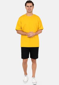 T-shirt en coton jaune à manches courtes, col rond. Associée à un short noir et des baskets blanches. Design simple, sans motifs visibles.