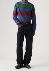 Långärmad randig polo i grön, blå, vinröd och mörk lila med denimkrage. Svarta vida byxor och svarta skor fulländar outfiten.