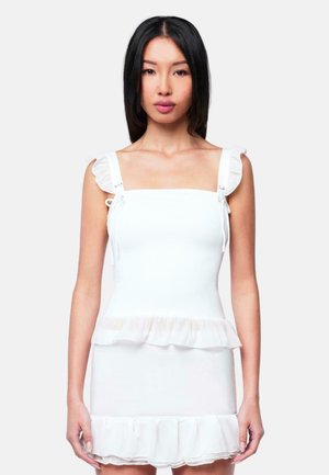 Donna con lunghi capelli neri che indossa un vestito bianco senza maniche con spalline a volant e orlo arricciato, in piedi contro uno sfondo semplice.