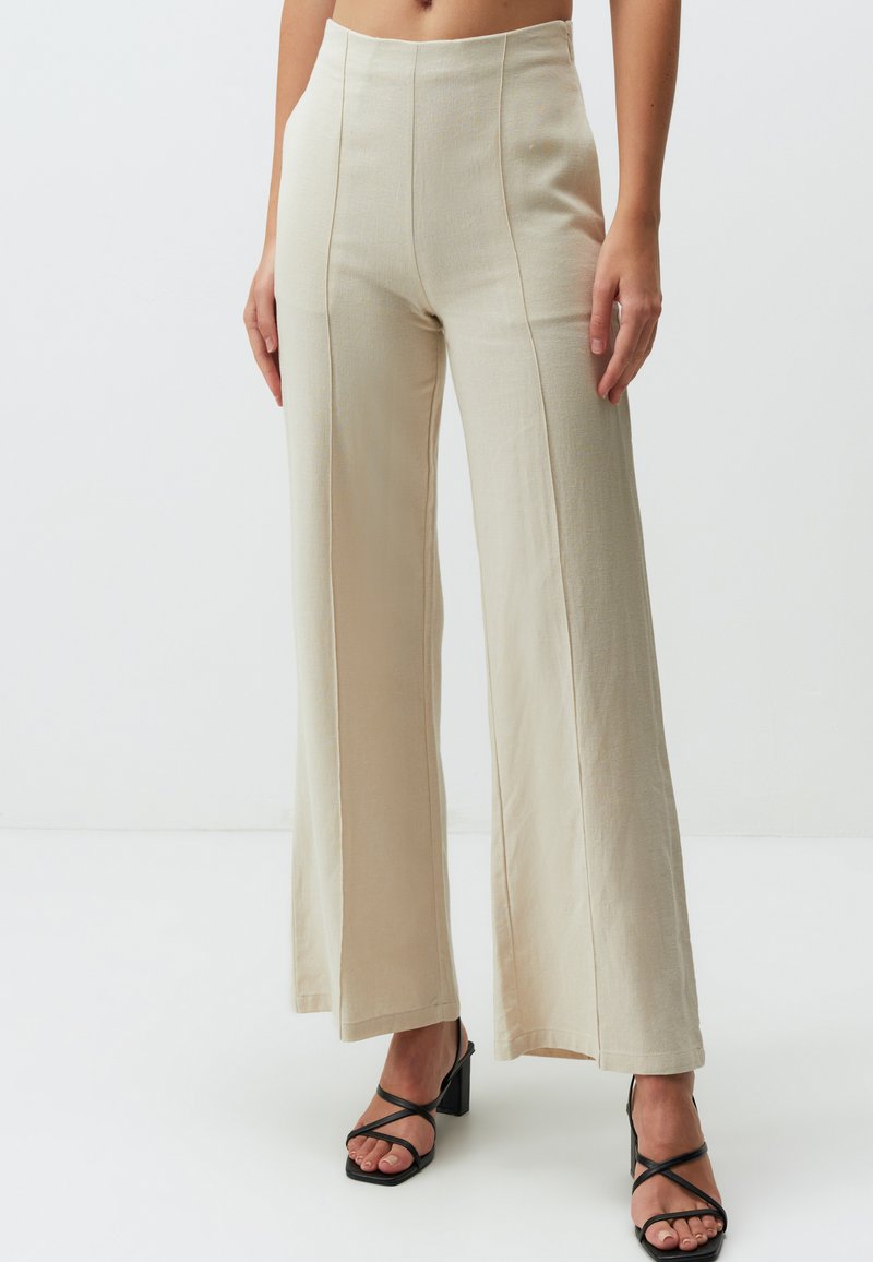 Pantalons larges beige à taille haute, avec des détails de couture visibles. Assortis à des sandales à talons bloc noires à lanières.