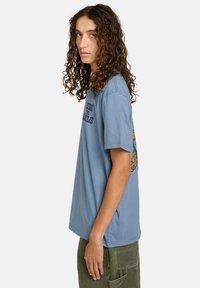 Ung person med langt krøllet hår, iført en blå grafisk t-shirt og grønne cargo-bukser, står sidelæns mod en hvid baggrund.