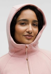 Svijetlo ružičasta zip-up hoodie s visokim ovratnikom, glatkom teksturom i naglasnim šavovima. Ima zlatni patentni zatvarač i podesivi kapuljaču.