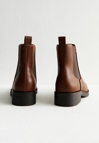 Botas de tobillo de piel marrón con paneles elásticos en los laterales, punta redonda, tacón pequeño y lengüeta en la parte trasera. Textura suave y diseño minimalista.