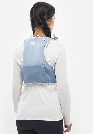 Femme aux cheveux tressés portant un gilet d'hydratation bleu et une chemise blanche à manches longues, vue de dos, montrant l'arrière du gilet et une bouteille d'eau.