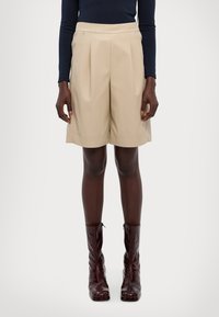 Beige plisserte short med skreddersydd design, laget av glatt stoff, brukt med glinsende burgunder ankelboots. Rett ben, midt på låret lengde.