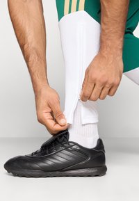 Melna sporta kurpe ar teksturētu virsmu; balta zeķe daļēji nosegta ar zaļu sporta legingu ar zeltainām akcentu svītrām, demonstrējot rāvējslēdzēja funkcionālās īpašības.