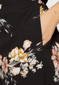 Tissu noir avec un motif floral dans des nuances de rose, jaune et blanc. Une poche latérale est visible, améliorant la fonctionnalité du vêtement.